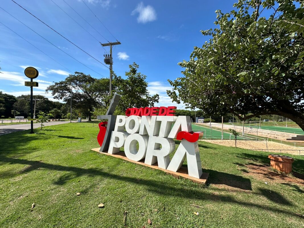Letrero con el nombre de Ponta Porã, en letras blancas y rojas, sobre el césped del Horto Florestal, con una cancha de césped sintético cercada al fondo bajo cielo azul