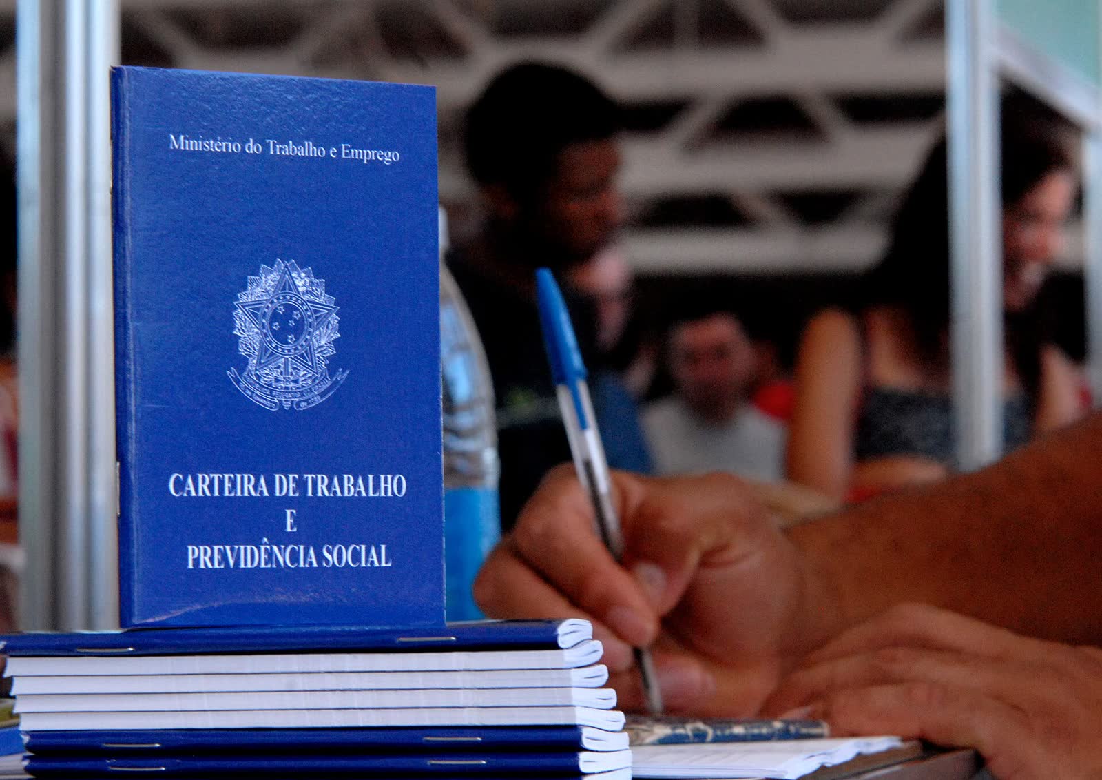 Carteira de Trabalho e Previdência Social azul sobre uma pilha de carteiras, com a mão de uma pessoa preenchendo um formulário ao lado e pessoas desfocadas ao fundo