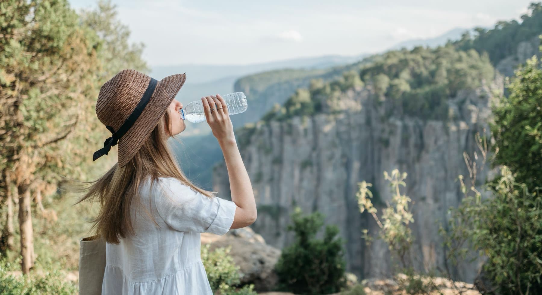 Mujer con sombrero de paja y vestido blanco toma agua de una botella al aire libre, frente a un paredón de piedra cubierto de monte