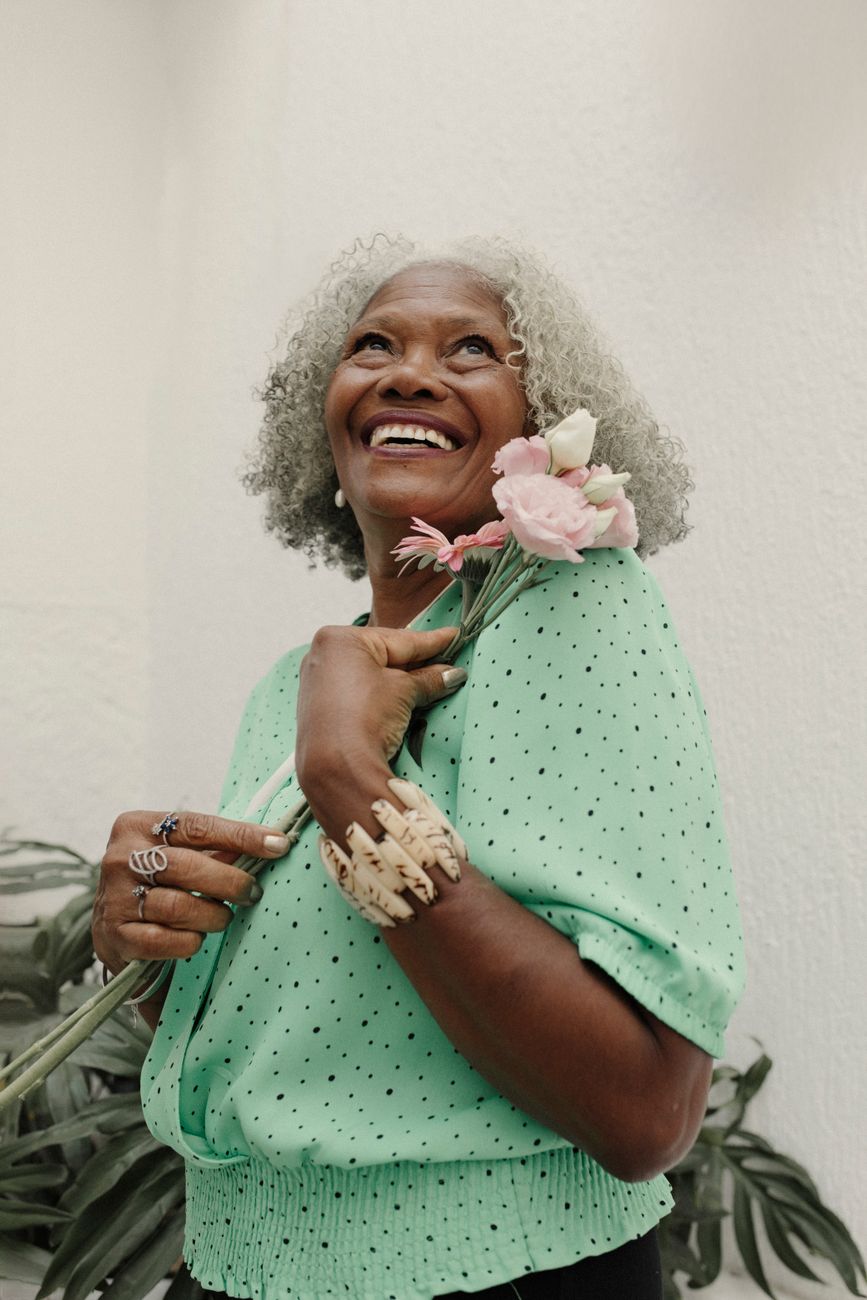 Mulher idosa de cabelo grisalho sorri segurando um ramo de flores cor de rosa