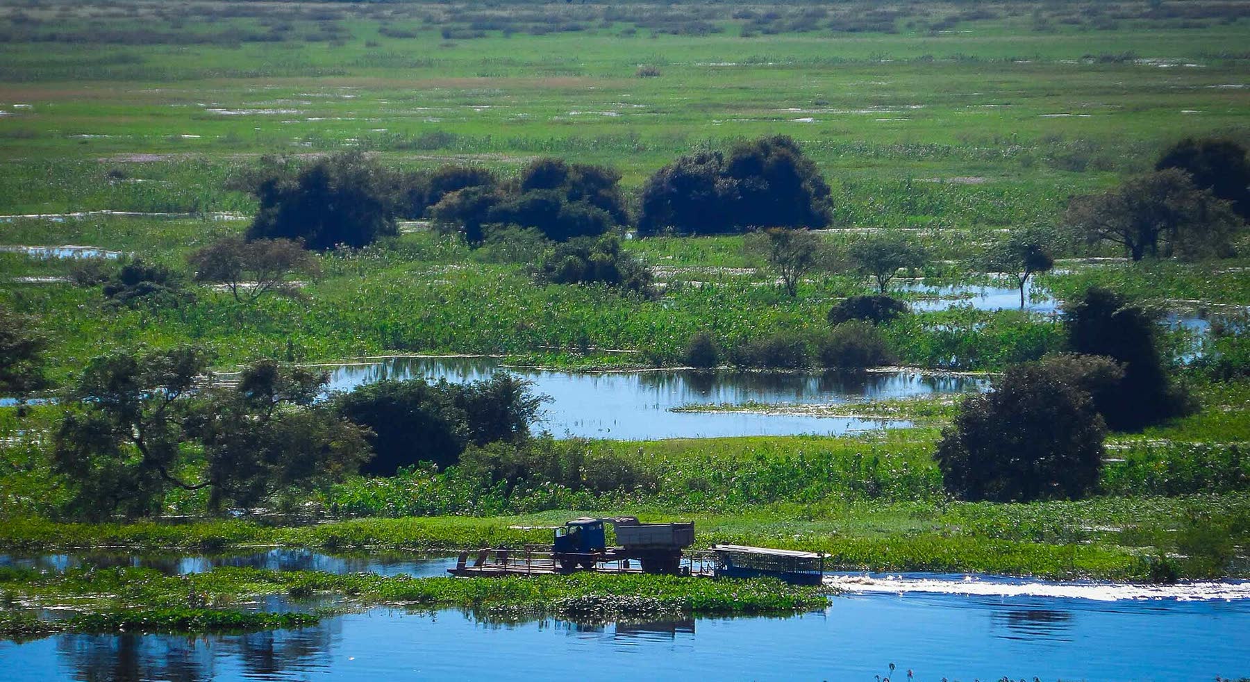 Paisagem alagada do Pantanal vista de cima, com vegetação verde, poças d'água e uma balsa pequena atravessando o rio com um caminhão a bordo.