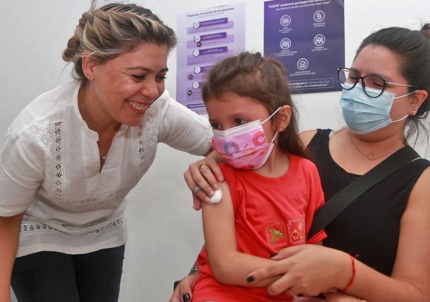 Menina de máscara, no colo de uma mulher, com algodão no braço depois da vacina, enquanto uma profissional sorri ao lado