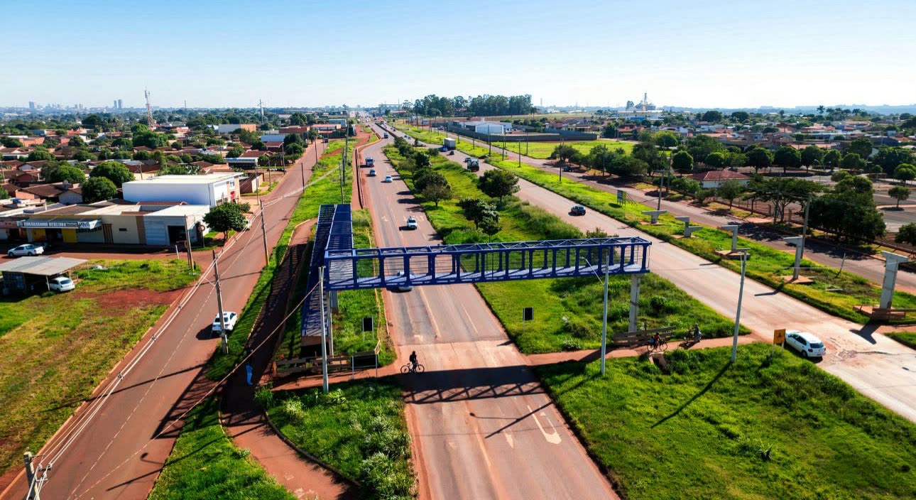 Pasarela peatonal azul sobre la ruta MS-156, en el trevo del DOF, en Dourados, vista desde arriba con barrios alrededor y ciclistas en la calzada