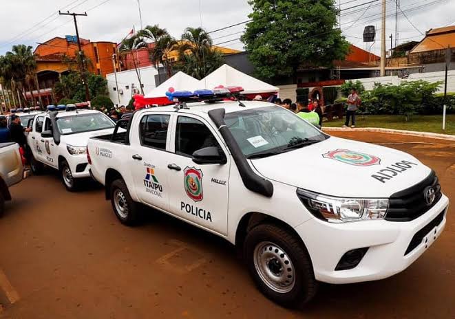 Fila de camionetes brancas da polícia estacionadas em uma rua de terra, com o escudo do Paraguai e a palavra Policía na porta e sinalizador azul e vermelho no teto
