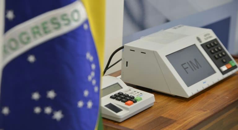 Urna eletrônica sobre uma mesa de madeira, com o teclado do mesário ao lado e uma bandeira do Brasil desfocada ao fundo, mostrando a mensagem FIM na tela.