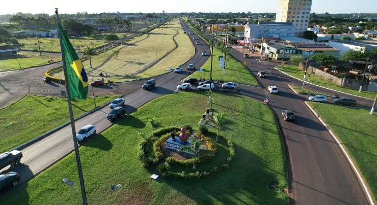 Vista aérea de uma rotatória com bandeira do Brasil hasteada, jardim central e ruas com carros em movimento, prédios ao fundo.