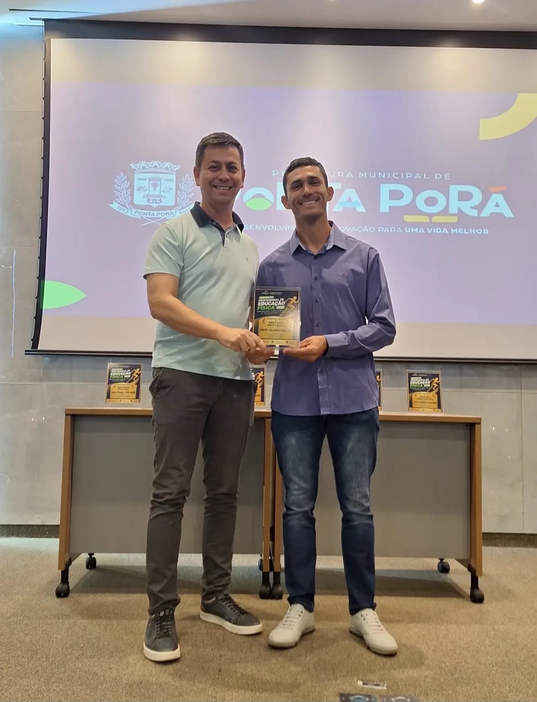 Fredy Miranda, de camisa azul, recibe una placa de homenaje de manos de otro hombre, frente a una pantalla con la marca de la Municipalidad de Ponta Porã