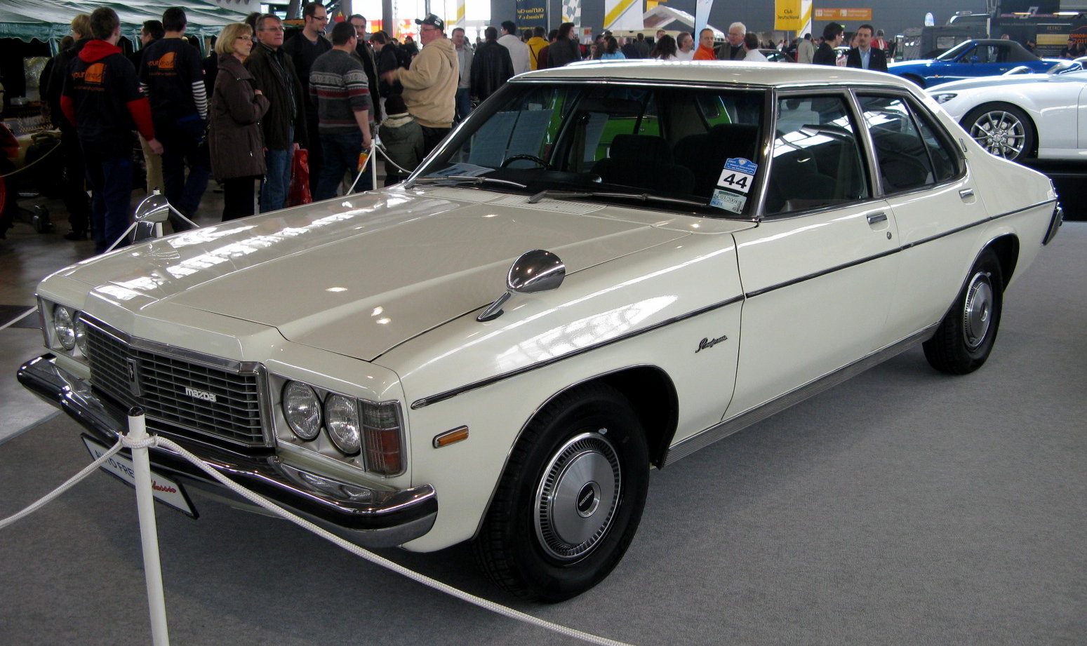 Sedã Mazda Roadpacer branco de 1979 exposto atrás de uma corda numa feira de carros antigos, com visitantes ao fundo