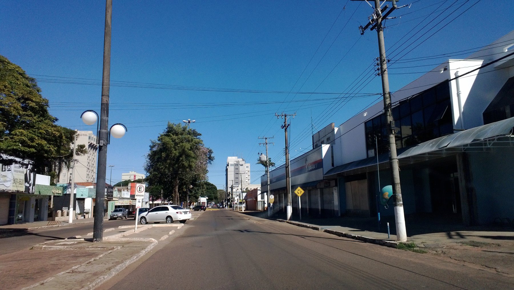 Avenida de Ponta Porã em dia de céu azul, com a rua quase vazia, postes de luz, árvores, lojas fechadas à direita e prédios ao fundo