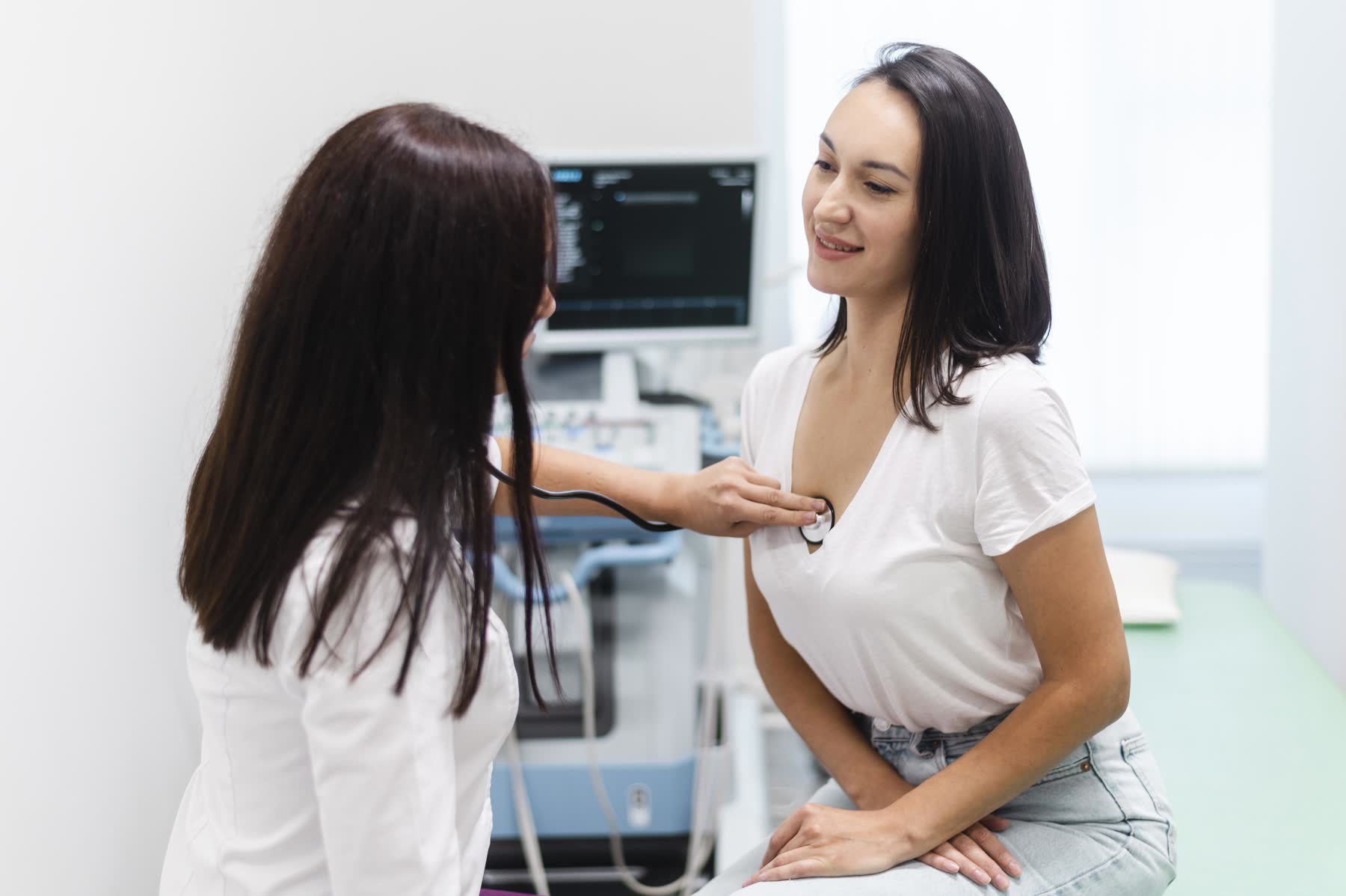 Médica de jaleco branco encosta o estetoscópio no peito de uma paciente sorridente, em consultório com aparelho de ultrassom ao fundo