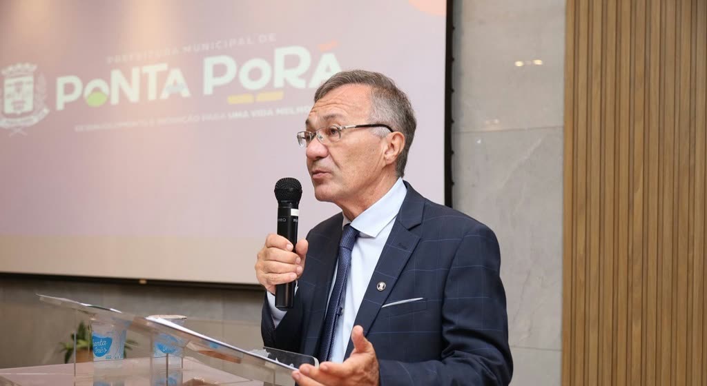 Desembargador Luiz Tadeu Barbosa Silva fala ao microfone num púlpito, com painel da Prefeitura de Ponta Porã ao fundo