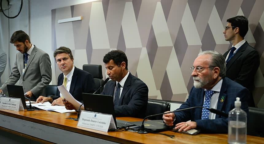 Mesa da comissão mista da MP do Enamed em 15 de outubro, com homens de terno sentados diante de papéis e plaquetas de identificação, e um deles de pé ao fundo.