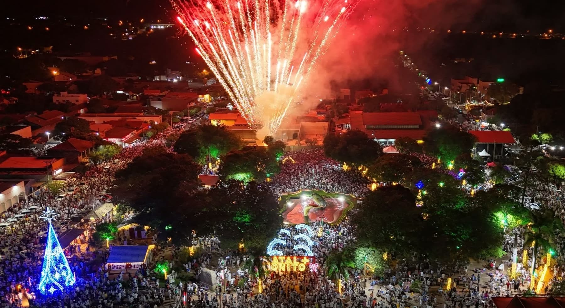 Vista aérea nocturna de la Plaza da Liberdade en Bonito llena de gente vestida de blanco, con fuegos artificiales rojos estallando sobre la multitud en un réveillon anterior