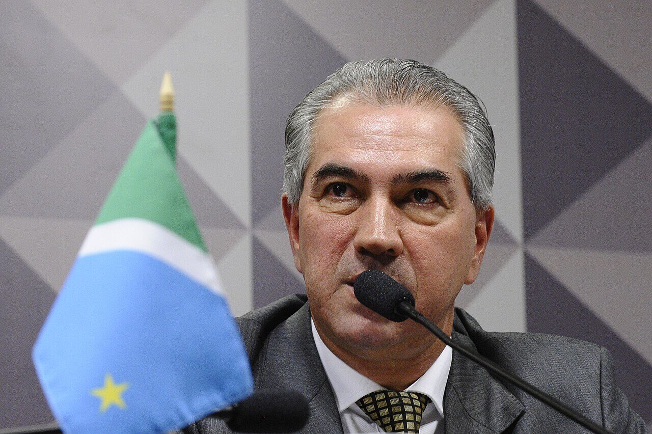 Reinaldo Azambuja fala ao microfone numa mesa do Senado, de terno cinza, com a bandeira de Mato Grosso do Sul em primeiro plano