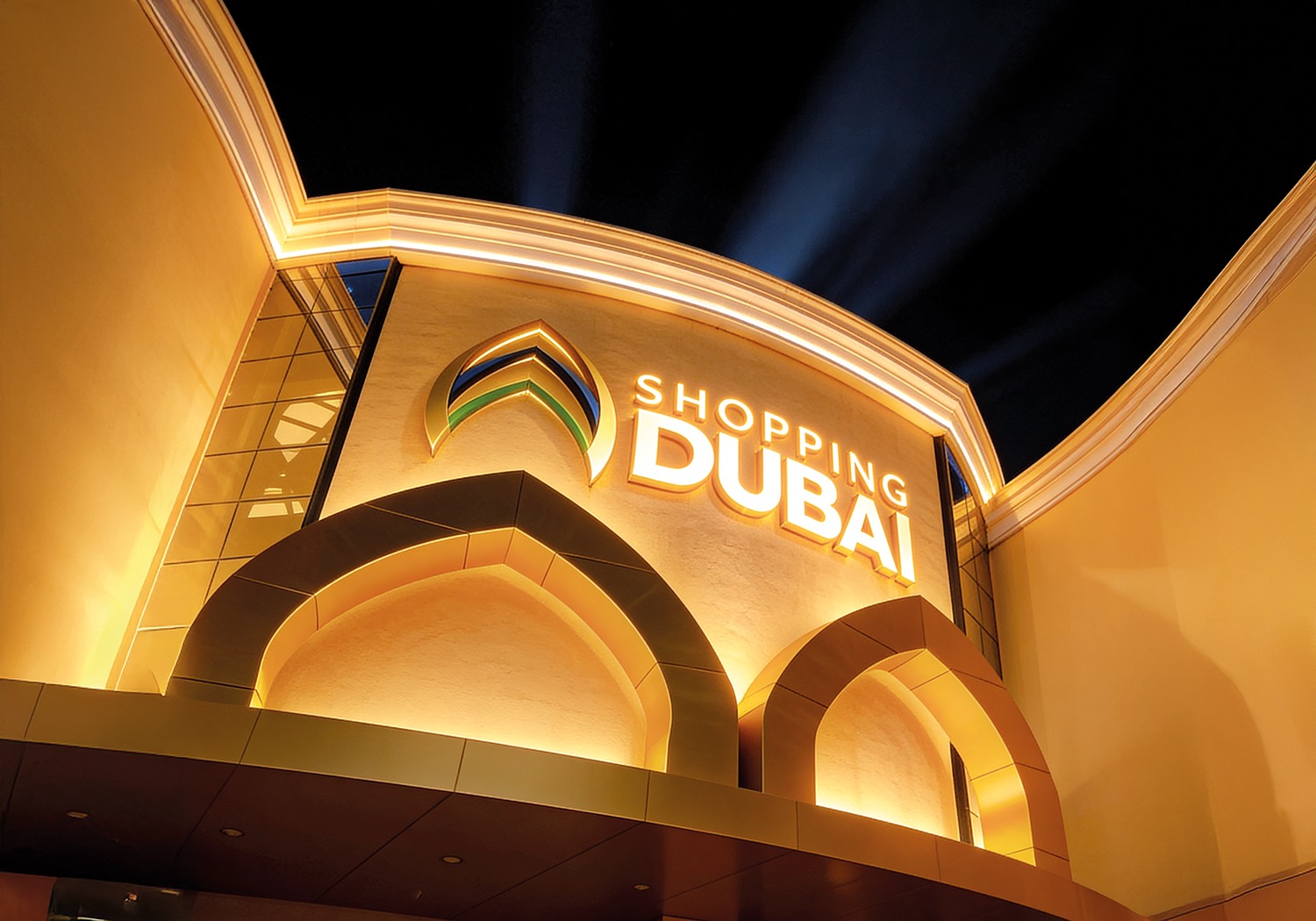 Fachada iluminada do Shopping Dubai à noite, com arcos e o letreiro do centro comercial
