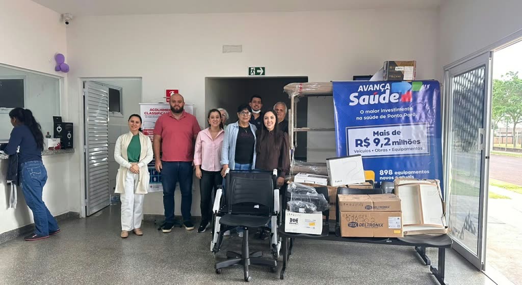 Equipe da saúde posa ao lado de caixas de equipamentos novos e de uma cadeira de rodas, diante de um banner do programa Avança Saúde