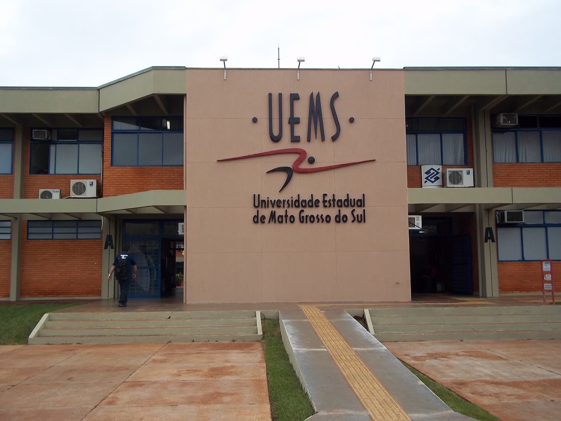 Fachada do prédio da UEMS em Dourados, com o nome da universidade em letras escuras sobre a parede rosada e um estudante entrando pela porta lateral