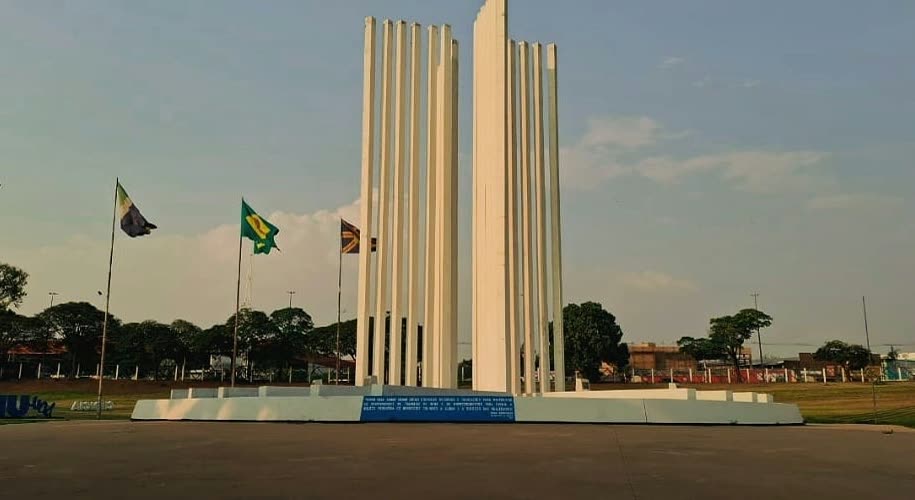 Monumento de colunas brancas verticais na entrada de um campus universitário, com bandeiras hasteadas ao redor.