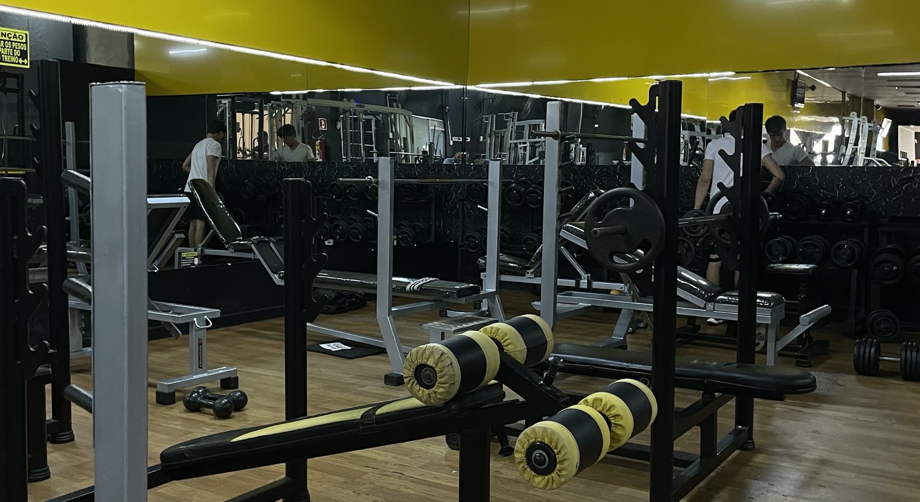 Sala de musculación con bancos y barras negras y amarillas frente a una pared con espejos