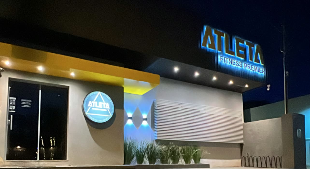 Fachada iluminada de noche con el letrero Atleta Fitness Premier en lo alto y el logotipo redondo encendido en la pared