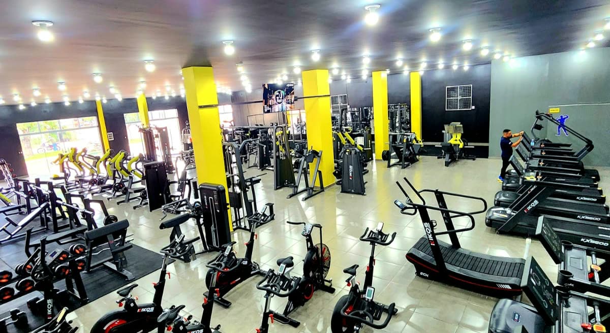 Amplio salón de gimnasio con cintas de correr, bicicletas y aparatos entre columnas amarillas