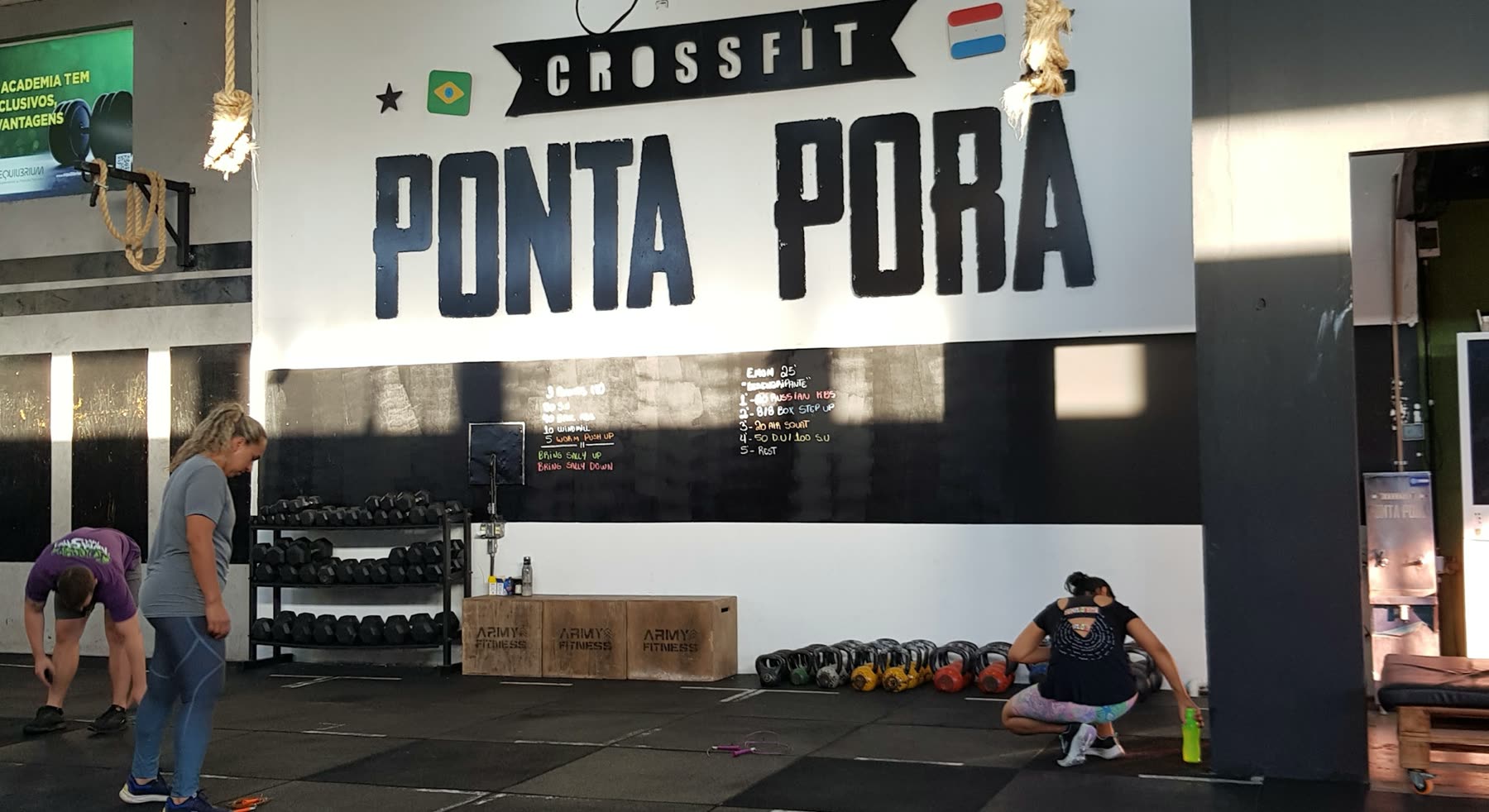Pared pintada con CrossFit Ponta Porã, pizarra de entrenamiento, cajones de madera y kettlebells, con personas entrenando en la colchoneta