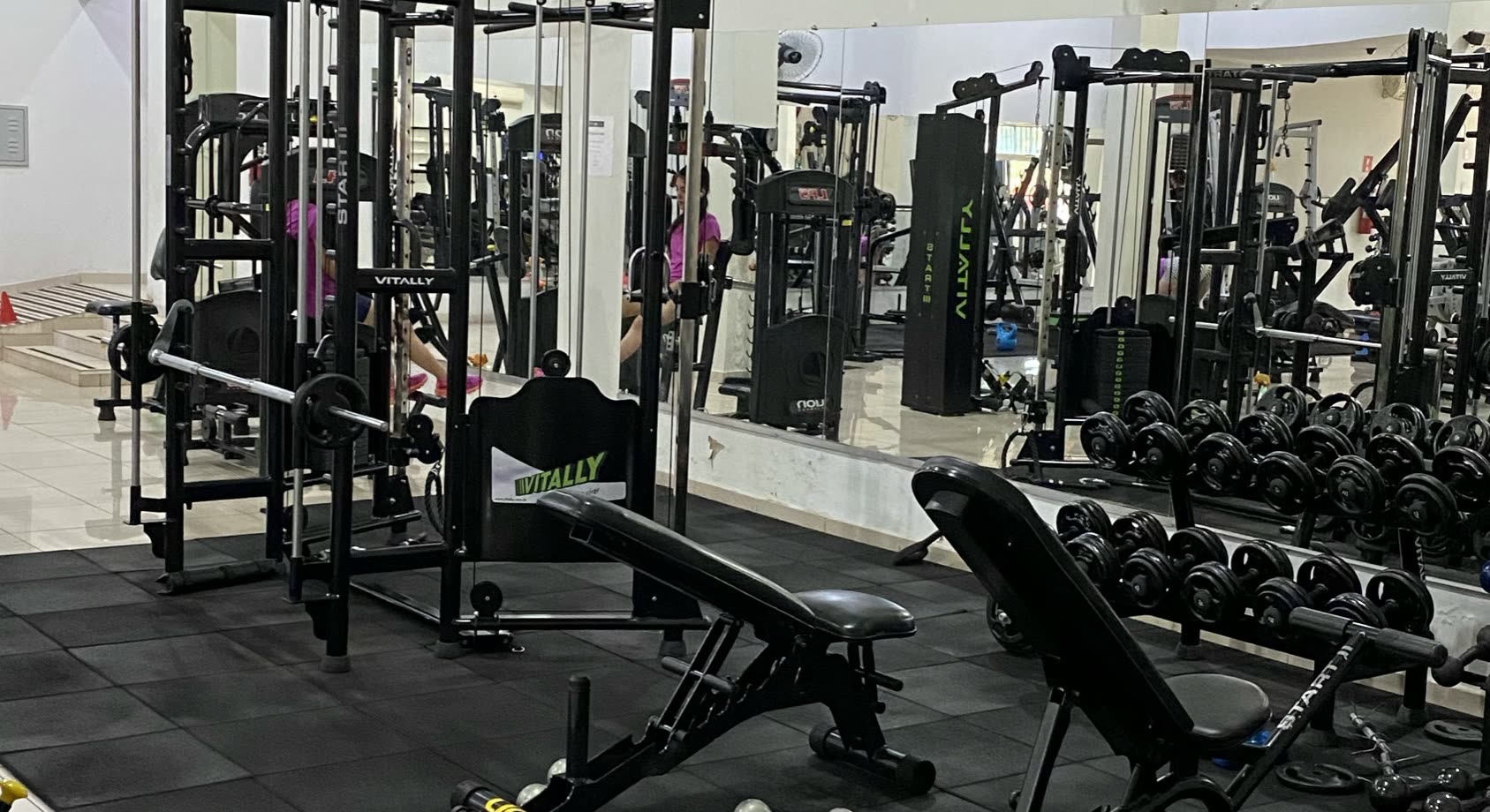 Estación de poleas, bancos y soporte de mancuernas frente a la pared con espejos de un gimnasio