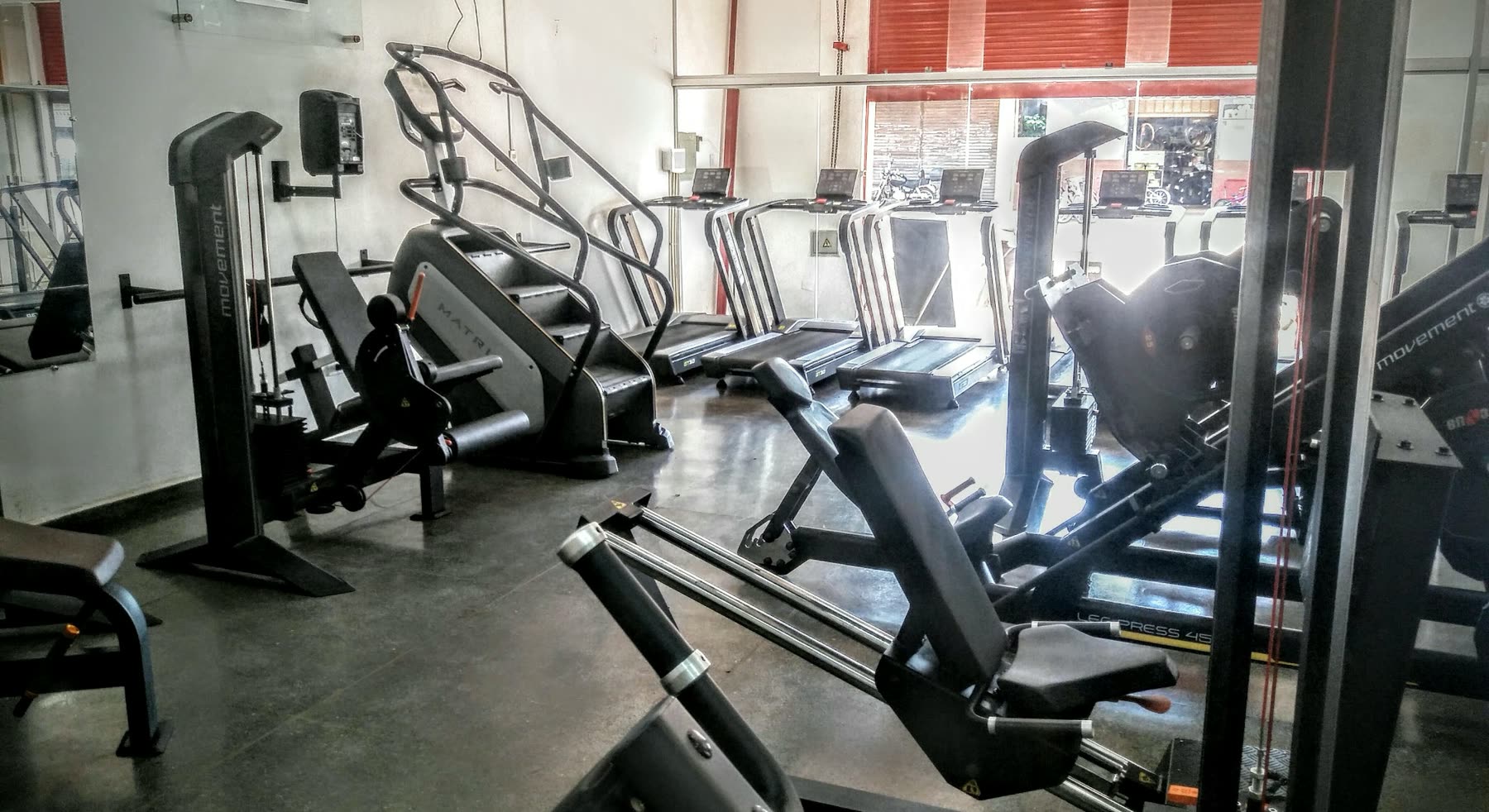 Cintas de correr en fila junto a aparatos de prensa de piernas y poleas, sobre el piso oscuro de un gimnasio