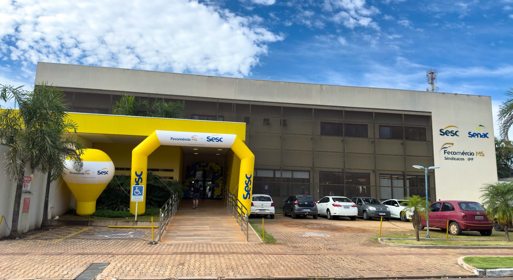 Edificio beige del Sesc con arco inflable amarillo en la entrada, rampa de acceso y autos estacionados enfrente