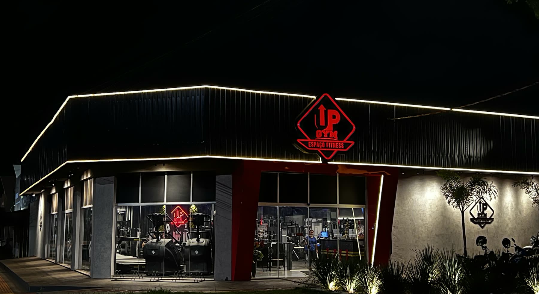 Fachada oscura de noche, bordeada por una tira de luz, con el logotipo rojo UP Espaço Fitness encendido sobre la entrada de vidrio