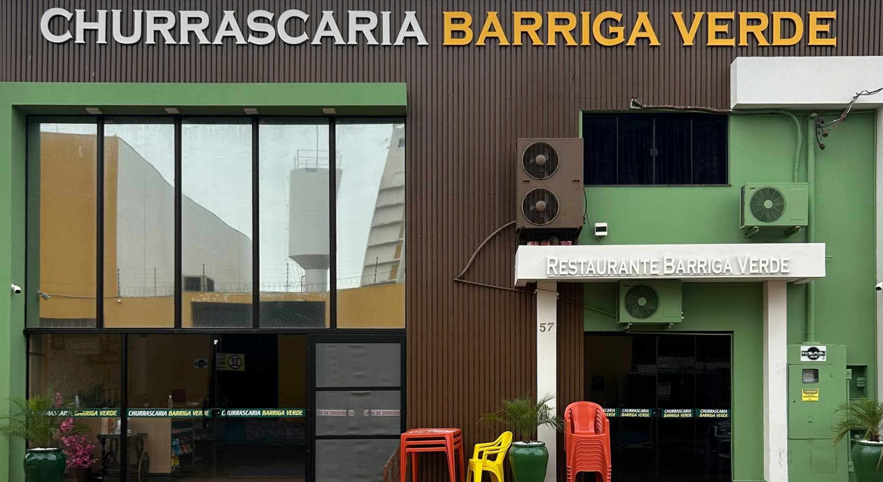 Fachada verde con el letrero Churrascaria Barriga Verde, puerta de vidrio y sillas plásticas de colores en la vereda