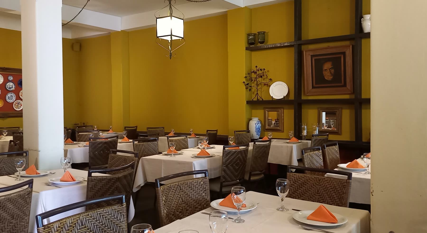 Salón de paredes amarillas con mesas puestas, manteles blancos, servilletas naranjas y cuadros en la pared