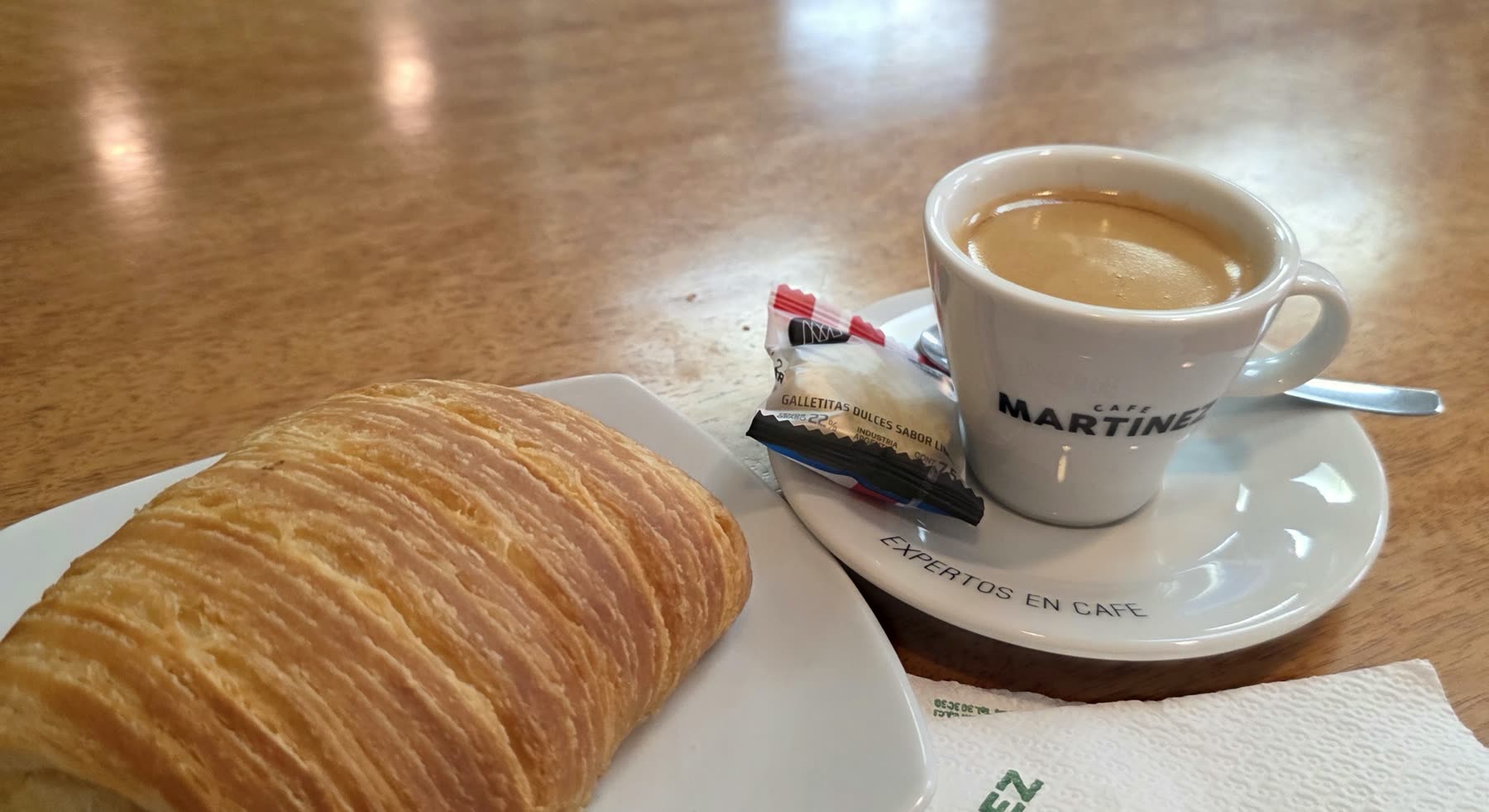 Taza blanca de café con la marca Martínez, junto a un croissant en el plato, sobre mesa de madera clara