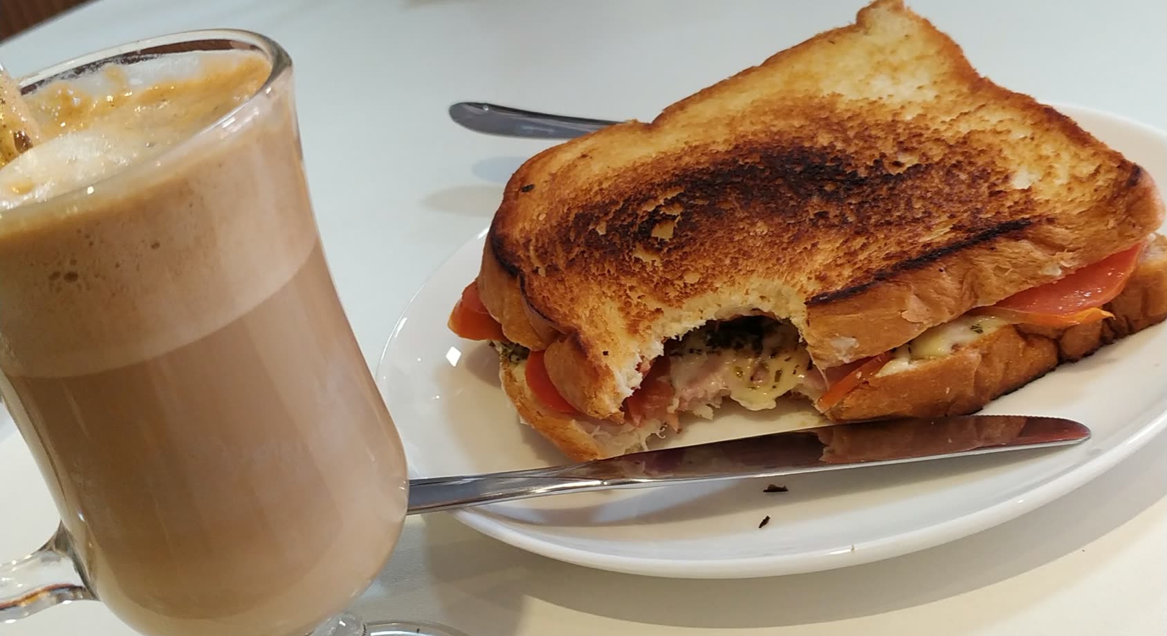 Vaso alto de café con leche junto a un sándwich tostado con tomate y jamón en el plato