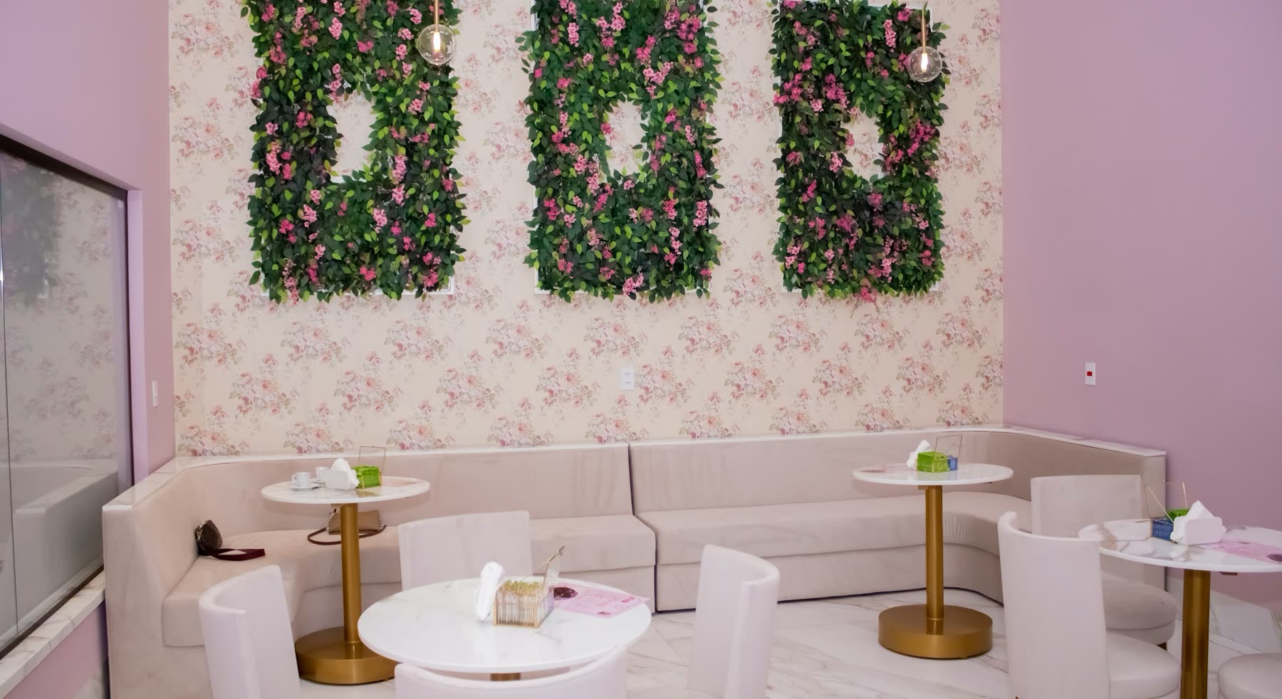 Salón en tonos rosados con paneles de flores en la pared, sofá claro y mesas redondas de base dorada
