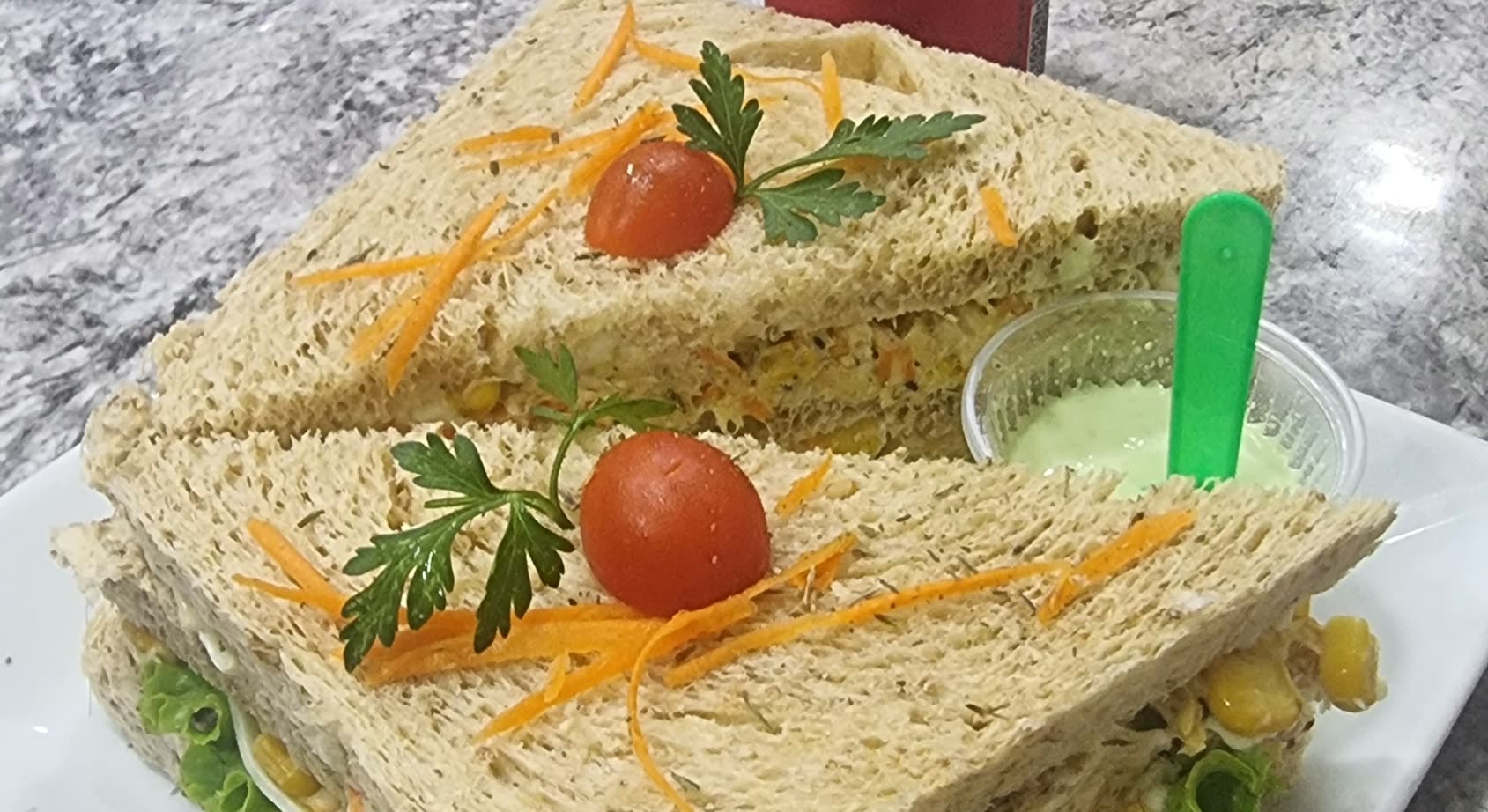 Dos sándwiches de pan integral cortados en triángulo, con zanahoria rallada, tomate cherry y perejil, y un pote de salsa verde con cuchara