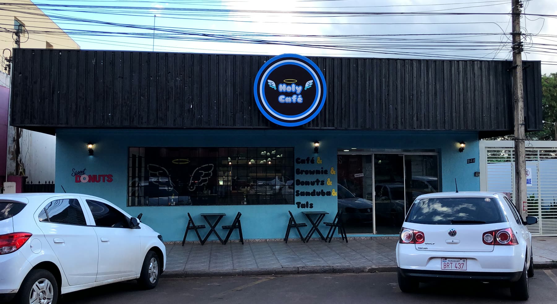 Fachada azul con letrero redondo de neón Holy Café, mesas altas en la vereda y autos estacionados enfrente