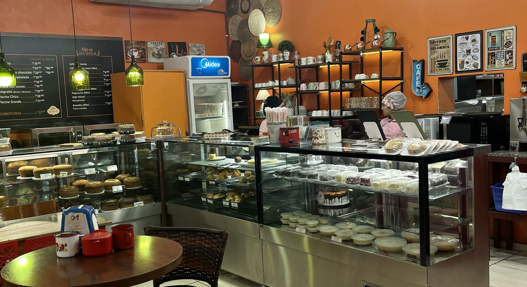 Salón de paredes naranjas con vitrinas refrigeradas llenas de tortas y dulces y el menú escrito en pizarra