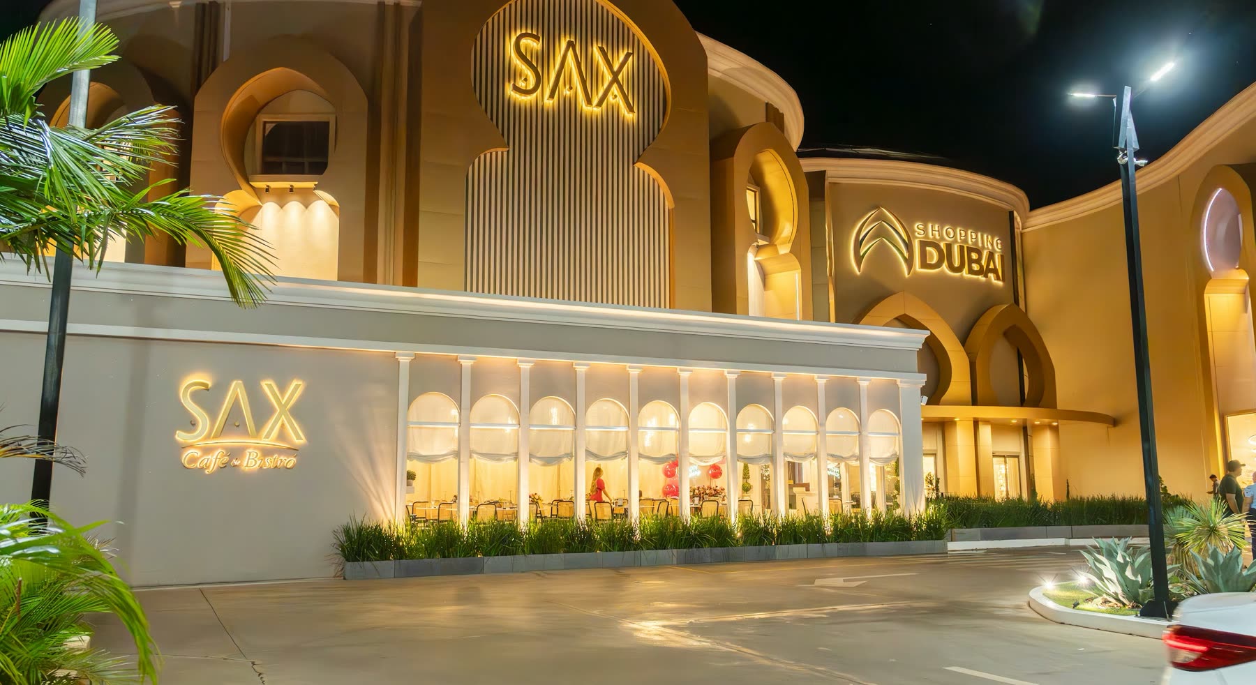 Fachada iluminada de noche con el letrero dorado SAX y, al lado, la entrada en arcos del Shopping Dubai
