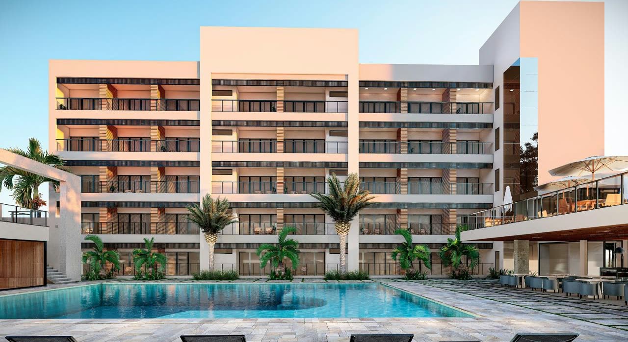 Edificio de hotel de cinco pisos con balcones, visto desde detrás de la piscina rodeada de palmeras y reposeras
