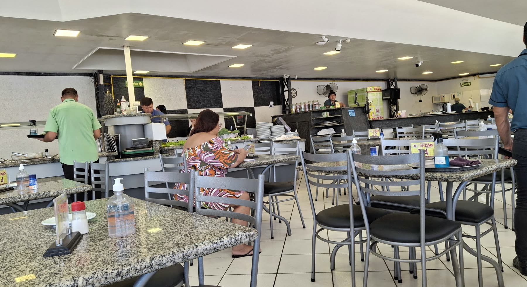 Salón de self-service con mesas de granito y sillas negras, y personas sirviéndose en el mostrador del bufé al fondo
