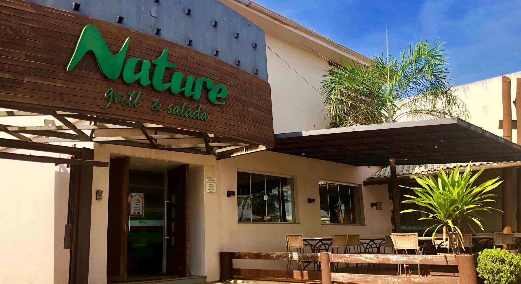Fachada con panel de madera y el letrero verde Nature grill & salada, con pérgola y mesas afuera