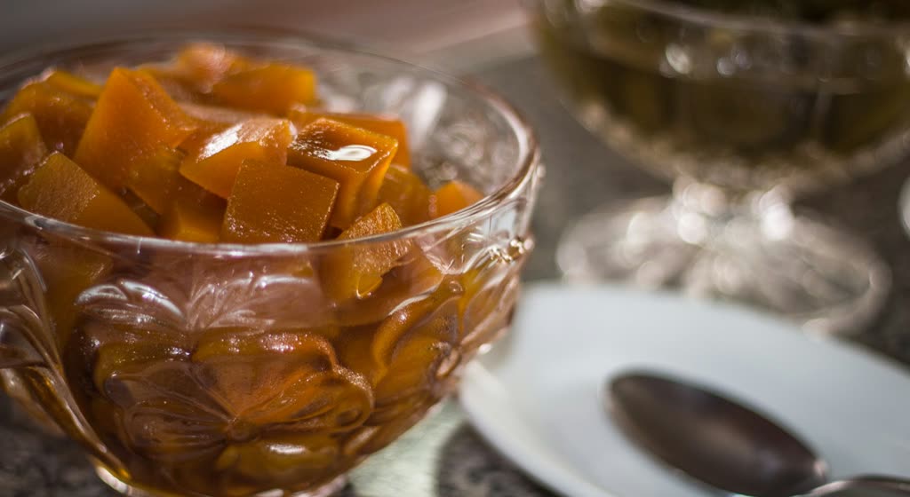 Copa de vidrio llena de cubos de dulce anaranjado en almíbar, con plato y cuchara al lado