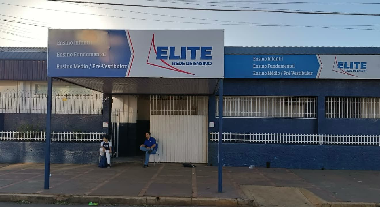Fachada azul y blanca con el letrero Elite Rede de Ensino que enumera educación inicial, primaria, media y preuniversitaria, con personas sentadas bajo la marquesina