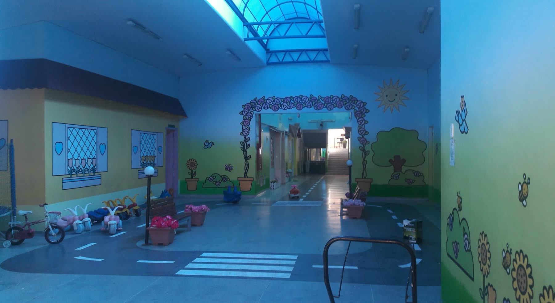 Patio cubierto por techo traslúcido azul, con paredes pintadas con dibujos infantiles, triciclos apoyados y una senda peatonal pintada en el piso