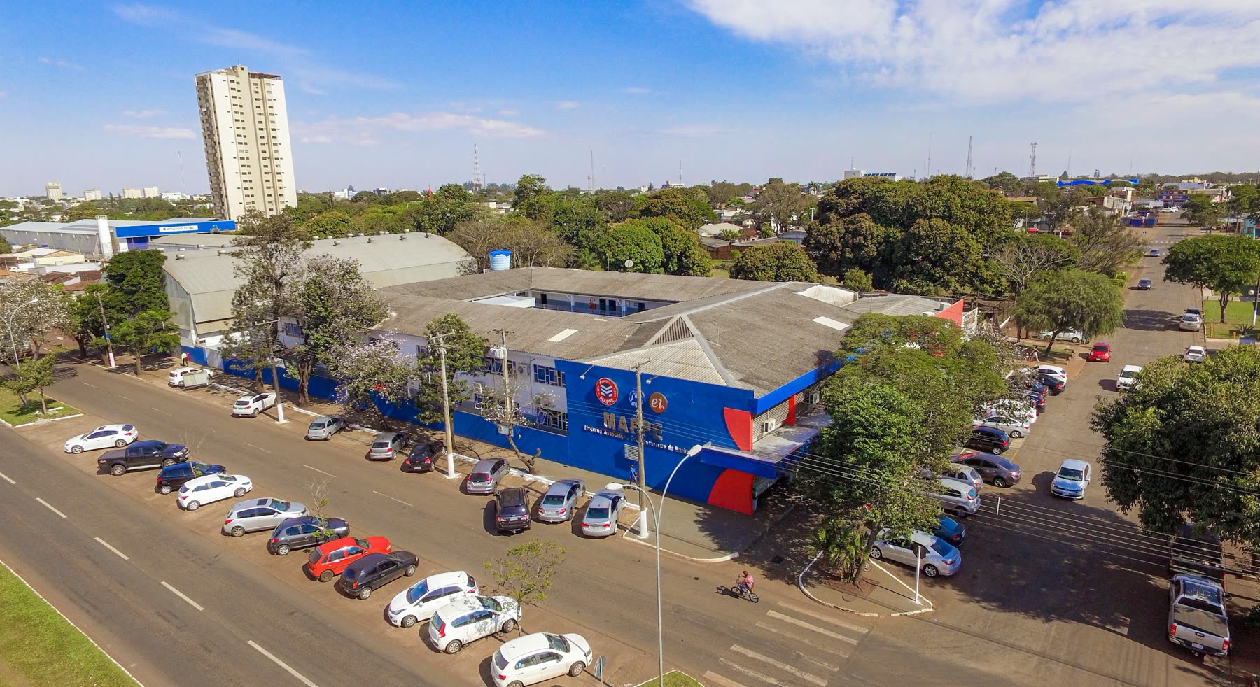 Vista aérea de un edificio escolar azul y blanco en la esquina, con autos estacionados a lo largo de la calle y árboles alrededor
