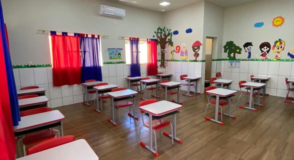 Aula vacía con pupitres blancos y rojos sobre piso de madera, cortinas de colores y pared decorada con un árbol y muñecos pintados