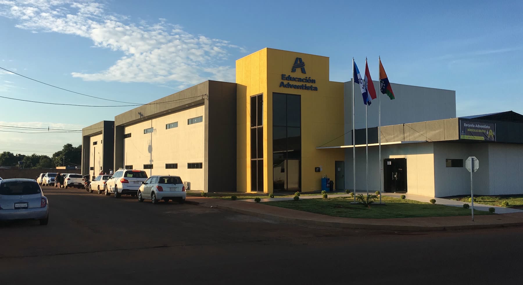Edificio amarillo y blanco con el logotipo de Educación Adventista en la torre, tres mástiles de bandera al costado y autos estacionados enfrente