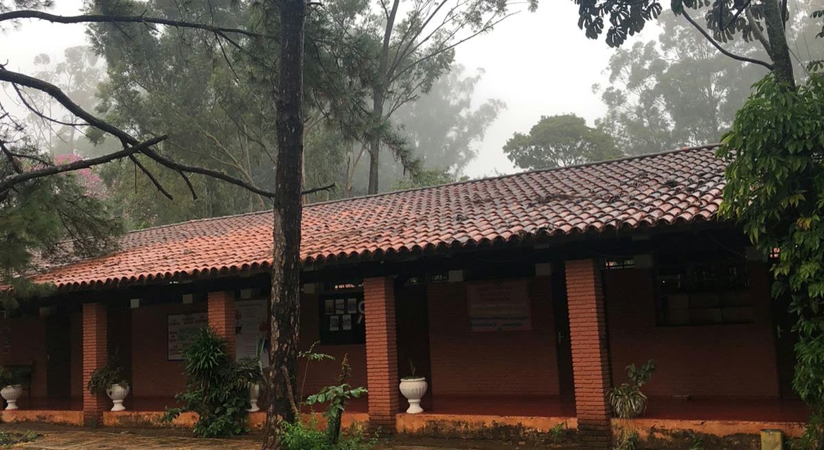 Construcción baja y alargada de techo de tejas y columnas de ladrillo, entre la neblina y árboles altos