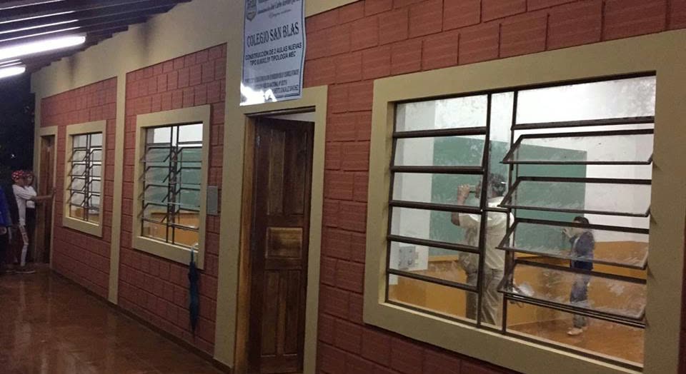 Pasillo de paredes de ladrillo con el cartel Colegio San Blas sobre la puerta de madera y ventanas enrejadas que dejan ver un aula en uso